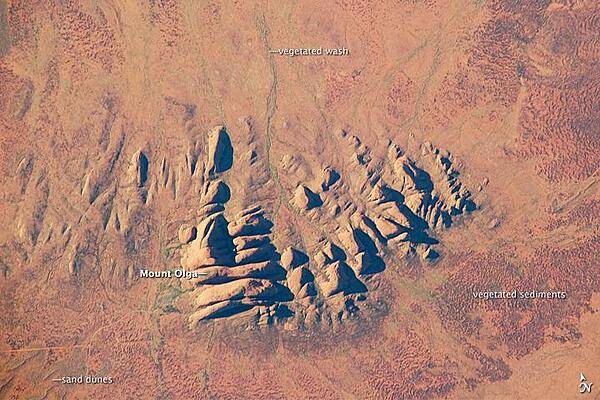 Located in the Northern Territory of Australia, Uluru-Kata Tjuta National Park hosts some of the world's most spectacular examples of inselbergs, or isolated mountains. The most famous of these inselbergs is Uluru (also known as Ayers Rock). An equally massive inselberg located approximately 30 km (20 mi) to the northwest is known as Kata Tjuta. Like Uluru, this is a sacred site to the native Anangu or Aboriginal people. An English-born explorer named the highest peak Mount Olga, with the entire grouping of rocks informally known as "the Olgas." Mount Olga has a peak elevation of 1,069 m (3,507 ft) above sea level, making it 206 m (676 ft) higher than Uluru.
In this astronaut photograph, afternoon sunlight highlights the rounded summits of Kata Tjuta against the surrounding sandy plains. Sand dunes are visible in the lower left, while in other areas (bottom and right) sediments washed from the rocks have been anchored by a variety of grasses and bushes adapted to the arid climate. Green vegetation in the ephemeral stream channels that drain Kata Tjuta (top center) provides colorful contrast with the red rocks and surrounding soils. Large gaps in the rocks (highlighted by shadows) are thought to be fractures that have been enlarged due to erosion. Image courtesy of NASA.