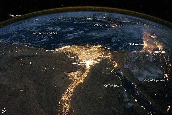 One of the fascinating aspects of viewing Earth at night is how well the lights show the distribution of people. In this view of Egypt, we see a population almost completely concentrated along the Nile Valley, just a small percentage of the country's land area.
The Nile River and its delta look like a brilliant, long-stemmed flower in this astronaut photograph of the southeastern Mediterranean Sea. The Cairo metropolitan area forms a particularly bright base of the flower. The smaller cities and towns within the Nile Delta tend to be hard to see amidst the dense agricultural vegetation during the day. However, these settled areas and the connecting roads between them become clearly visible at night. Likewise, infrastructure and urbanized regions along the Nile River also become apparent.
Another brightly lit region is visible along the eastern coastline of the Mediterranean, the Tel-Aviv metropolitan area in Israel (image right). To the east of Tel-Aviv lies Amman, Jordan. The two major water bodies that define the western and eastern coastlines of the Sinai Peninsula, the Gulf of Suez and the Gulf of Aqaba, are outlined by lights along their coastlines (image lower right). The city lights of Paphos, Limassol, Larnaca, and Nicosia are visible on the island of Cyprus (image top).
Scattered blue-grey clouds cover the Mediterranean Sea and the Sinai, while much of northeastern Africa is cloud-free. The thin yellow-brown band tracing the Earth's curvature at image top is airglow, a faint band of light emission that results from the interaction of atmospheric atoms and molecules with solar radiation at approximately 100 km (60 mi) altitude. Photo courtesy of NASA.