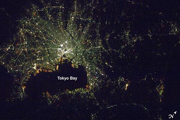 A nighttime view of Tokyo, Japan from the International Space Station. The heart of the city is brightest, with ribbons of lights radiating outward from the center along streets and railways. The regularly spaced bright spots along one of the ribbons heading almost due west out of the downtown area are train stations along a public transit route. The lights of Tokyo are a cooler blue-green color than in many other world cities. The color results from the more widespread use of mercury vapor lighting as opposed to sodium vapor lighting, which produces an orange-yellow light. Photo: NASA.