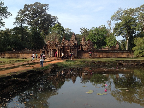 Banteay Srei is a 10th-century Cambodian temple dedicated to the Hindu god Shiva. The site lies 25 km (15 mi) north-east of the main group of temples at Angkor. Banteay Srei is built largely of red sandstone, a medium that lends itself to the elaborate decorative wall carvings that are still observable today. The buildings themselves are miniature in scale, unusually so when measured by the standards of Angkorian construction. These factors have made the temple extremely popular with tourists. This photo shows the entrance way and part of the moat that surrounds the temple complex.