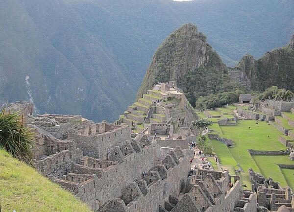 A section of housing is seen in front of Machu Picchu's Sacred Plaza, with the Intiwatana (Hitching Post of the Sun) above the plaza and the Western Urban Section to its right, where llamas roam.