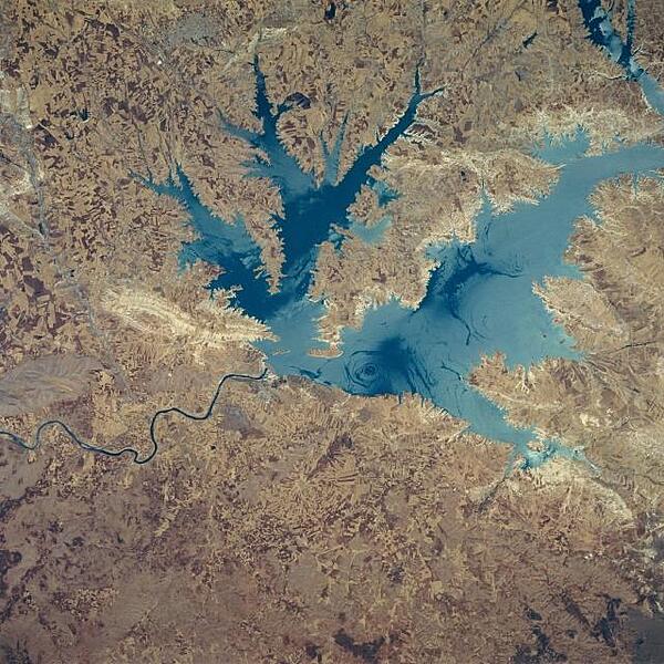 This northeast-looking view shows the Ataturk Reservoir and Dam, presently the largest man-made lake in Turkey. Located in south central Turkey on the Euphrates River, Ataturk Reservoir was completed in the early 1990's. The meandering Euphrates River (dark line) can be seen exiting the scene below the left center of the image. Smooth lake waters appear as dark blue, while wind-roughened waters appear light blue. The light area to the west of the dam (left center) is where rocks were quarried for the construction of the dam. Photo courtesy of NASA.