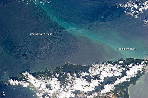 This space station photo of 18 January 2013, shows the north coast of Trinidad and a series of subtle, interacting arcs in the southeastern Caribbean Sea. These are known as "internal waves," the surface manifestation of slow waves that move tens of meters beneath the sea surface. Internal waves produce enough of an effect on the sea surface to be seen from space, but only where they are enhanced due to reflection of sunlight, or sunglint, back towards the space station. 
The image shows at least three sets of internal waves interacting. The most prominent set (image top left) shows a packet of several waves moving from the northwest due to the tidal flow towards the north coast of Trinidad. Two less prominent, younger sets can be seen further out to sea. A very broad set enters the view from the north and northeast, and interacts at image top center with the first set. All the internal waves are probably caused by the shelf break near Tobago (outside the image to top right). The shelf break is the step between shallow seas (around continents and islands) and the deep ocean. It is the line at which tides usually start to generate internal waves. 
Over the island of Trinidad, the heating of the land surface sets off the growth of cumulus clouds. Off the coast, a light blue northwest-southeast trending plume at image center is sediment embedded in the Equatorial Current (also known as the Guyana Current). The current is transporting material to the northwest - in almost the opposite direction of the internal waves. The current flows strongly from east to west around Trinidad, all the way from equatorial Africa, driven by year-round easterly winds. Seafarers in the vicinity of Trinidad are warned that the current - and its local reverse eddies - make navigation complicated and sometimes dangerous for smaller craft in these waters.
Astronauts also have observed internal waves in other parts of the world, such as San Francisco and the Straits of Gibraltar. Photo courtesy of NASA.