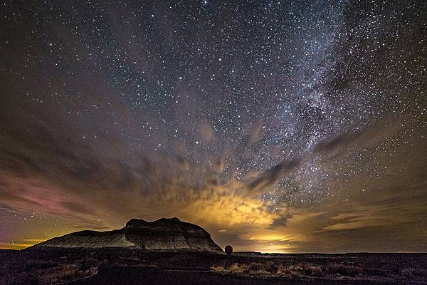 Stars of the Milky Way shine from behind lit clouds above The Battleship, a butte near Crystal Forest in Petrified Forest National Park, Arizona. Image courtesy of the US National Park Service/Jacob Holgerson.